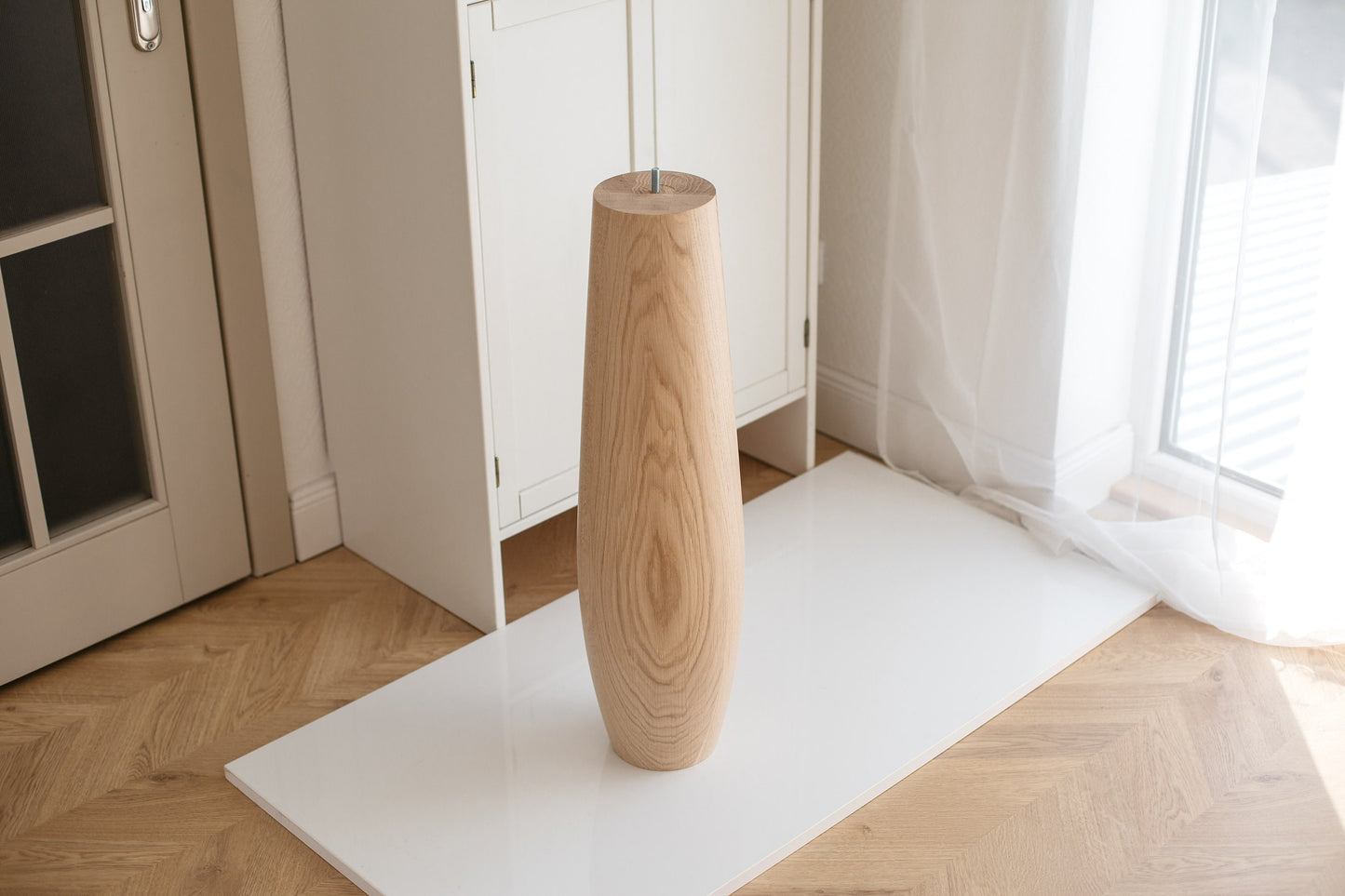 An oval wooden furniture leg, presumably a table leg, displayed against a white background with a light-colored floor.