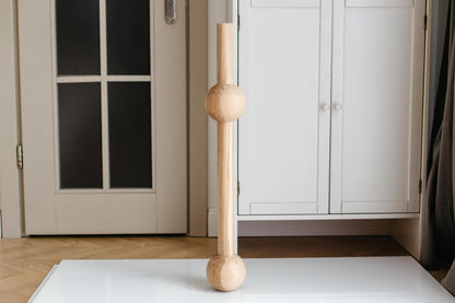 A single handmade wooden furniture leg with two spheres, placed on a white surface with a door and window in the background.