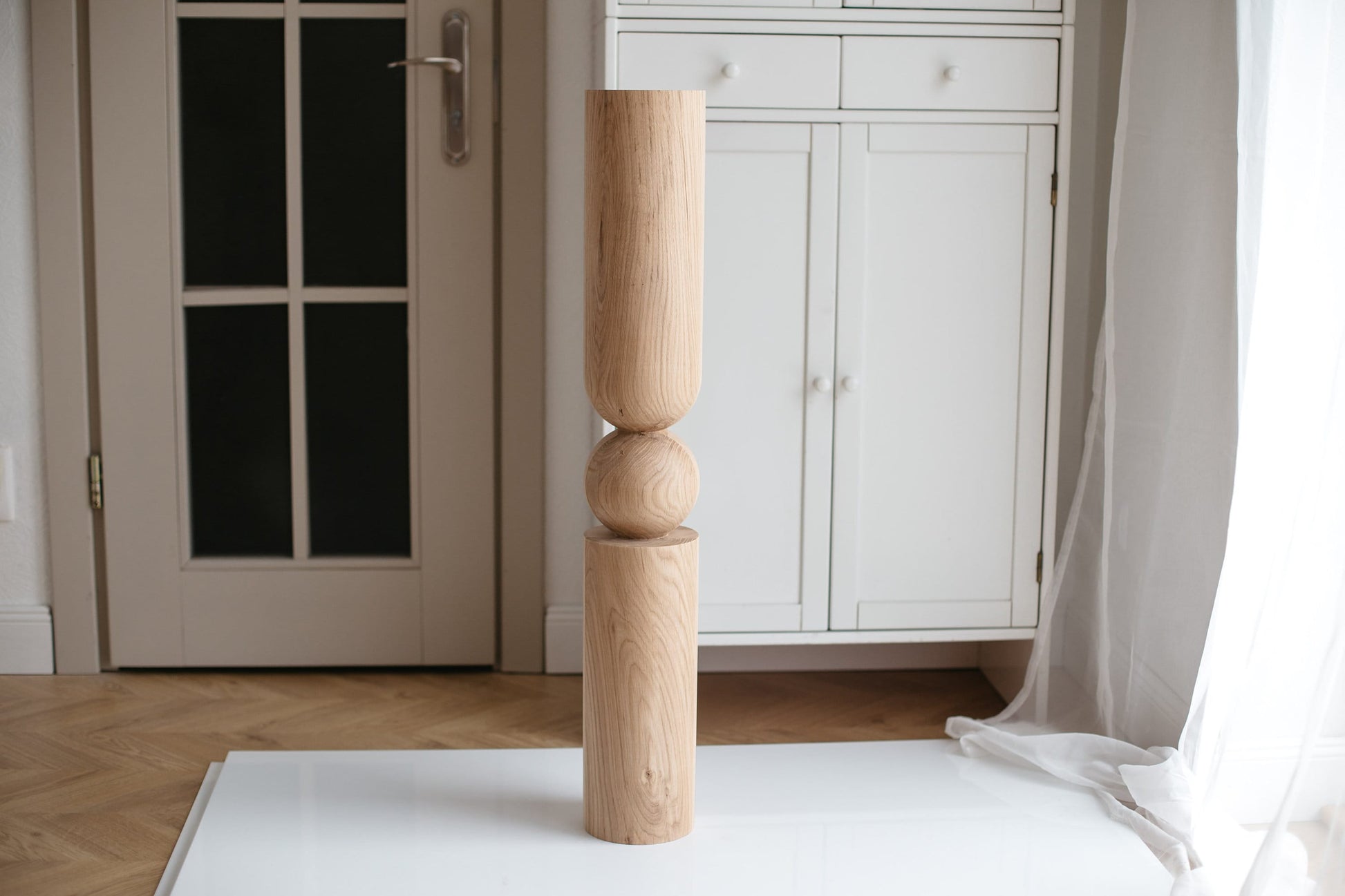 A single beige wooden furniture leg with a middle sphere standing upright on a white surface with a door and furniture in the background.