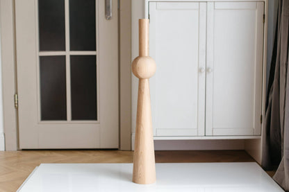 A single beige wooden furniture leg with a sphere on top stands against a white background, with a door and white furniture in the soft focus background.