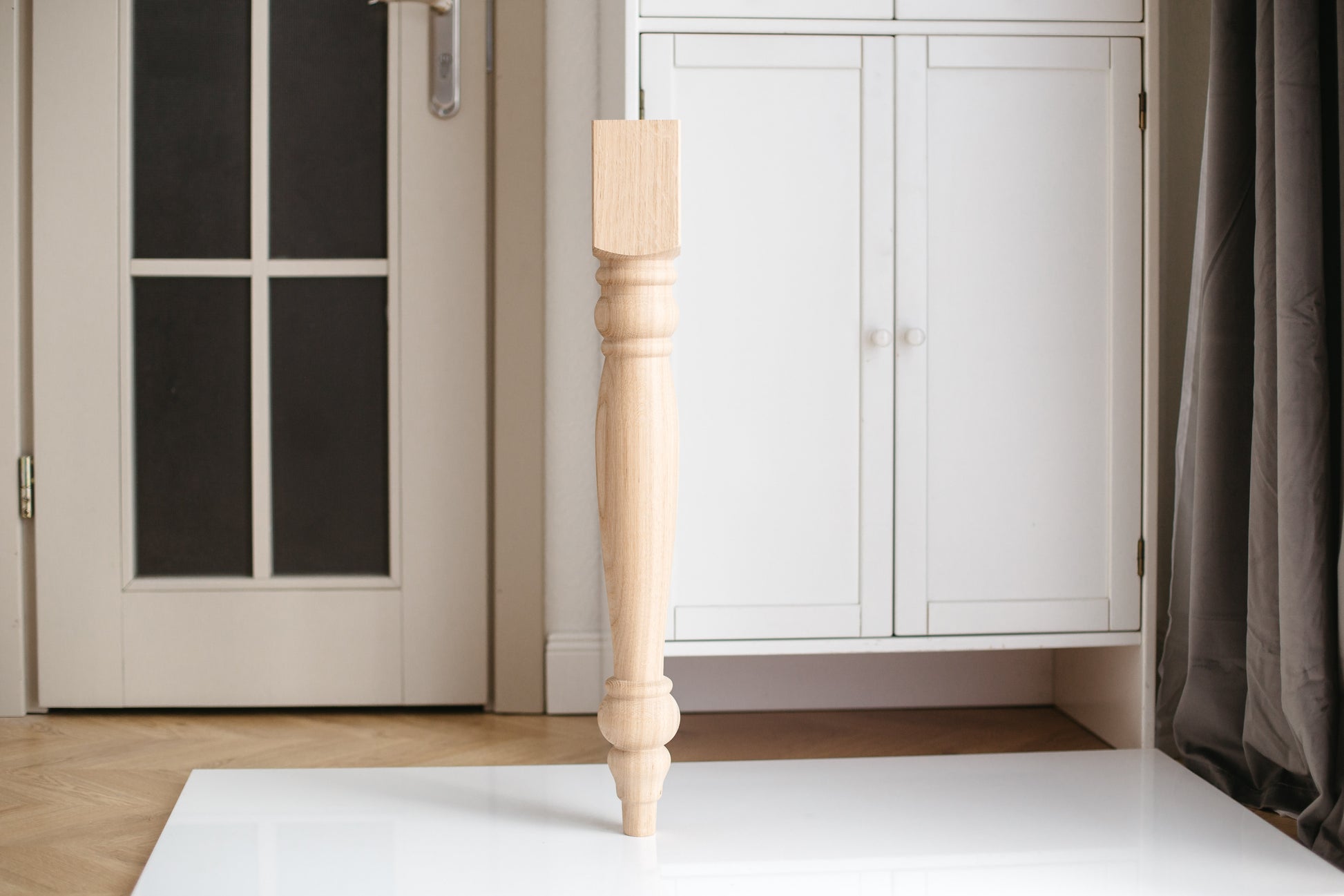 A single wooden furniture leg placed upright on a white surface with a door and cabinet in the background.