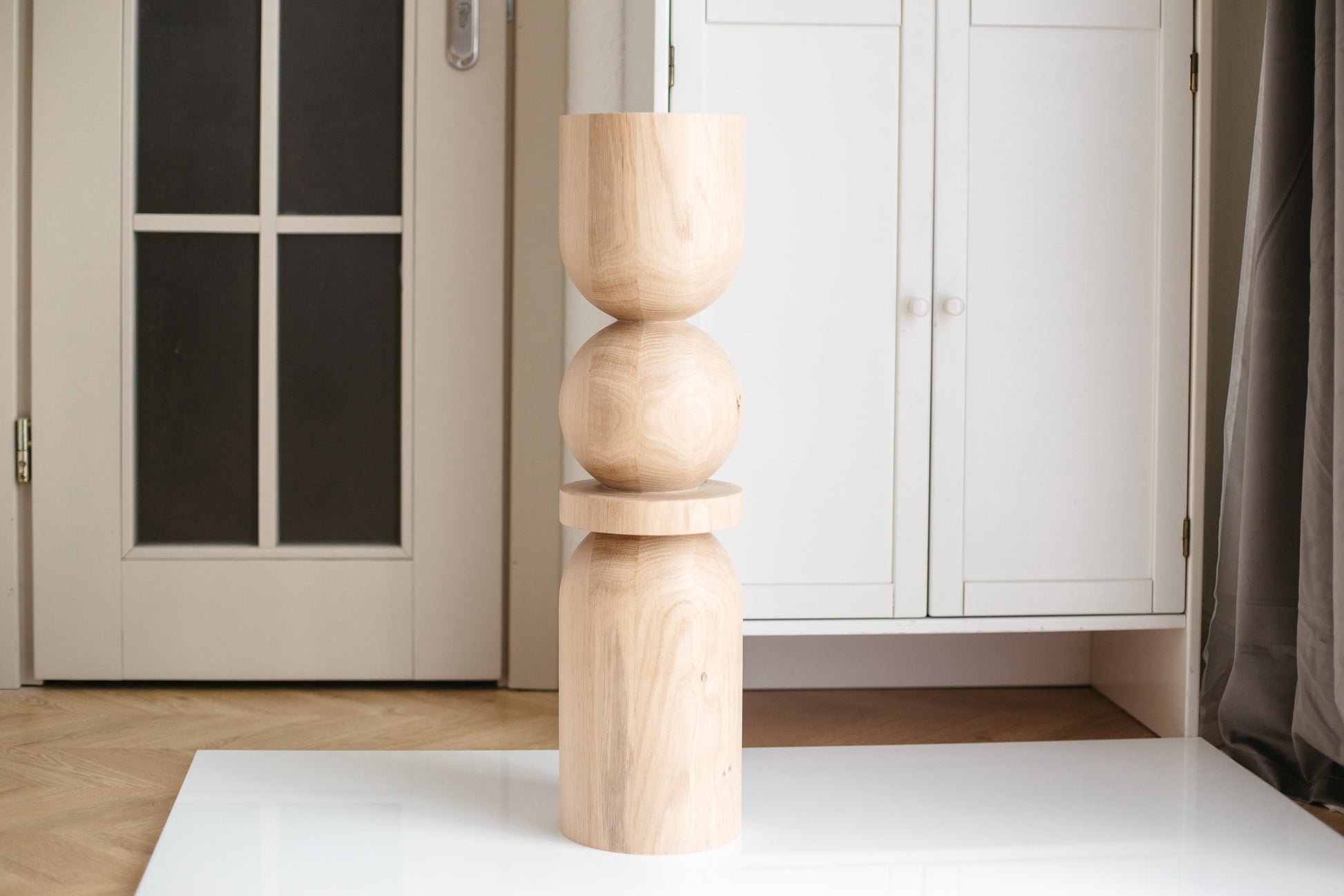 A single wooden furniture leg with a spherical design at the middle, placed on a white surface with a door and a wardrobe in the background.