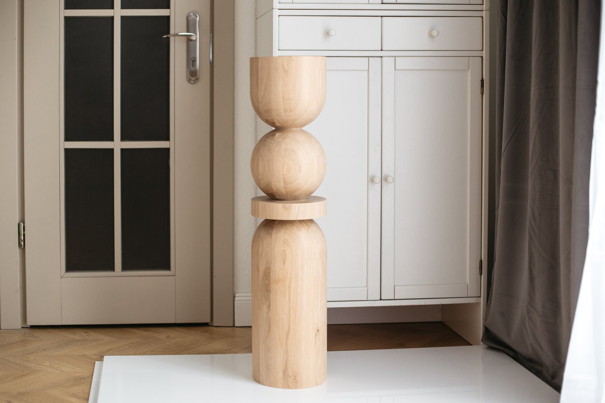 A single wooden furniture leg with a spherical design at the top, placed on a white surface, with a door and a white cabinet in the background.