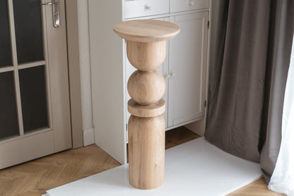 A cylindrical wooden pedestal table with a smooth finish, displayed in a room setting.