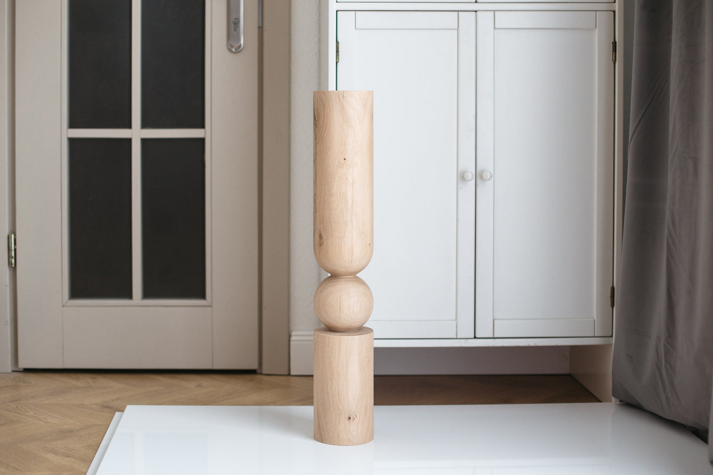 A single wooden furniture leg with a spherical middle section standing upright on a flat surface, with a door and a cabinet in the background.