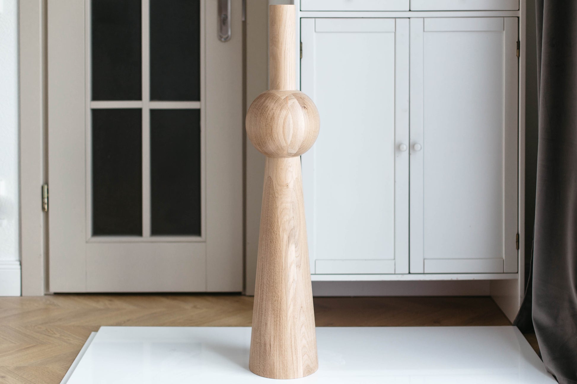 A light natural wood decorative object with a tapered base and a rounded middle section, standing on a white surface in an indoor room with white cabinets and a herringbone wood floor.