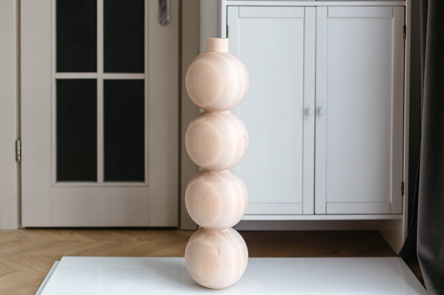 A tall, minimalist decorative vase made of four stacked, rounded wooden spheres in a light natural wood finish, standing on a white surface in an indoor room setting.