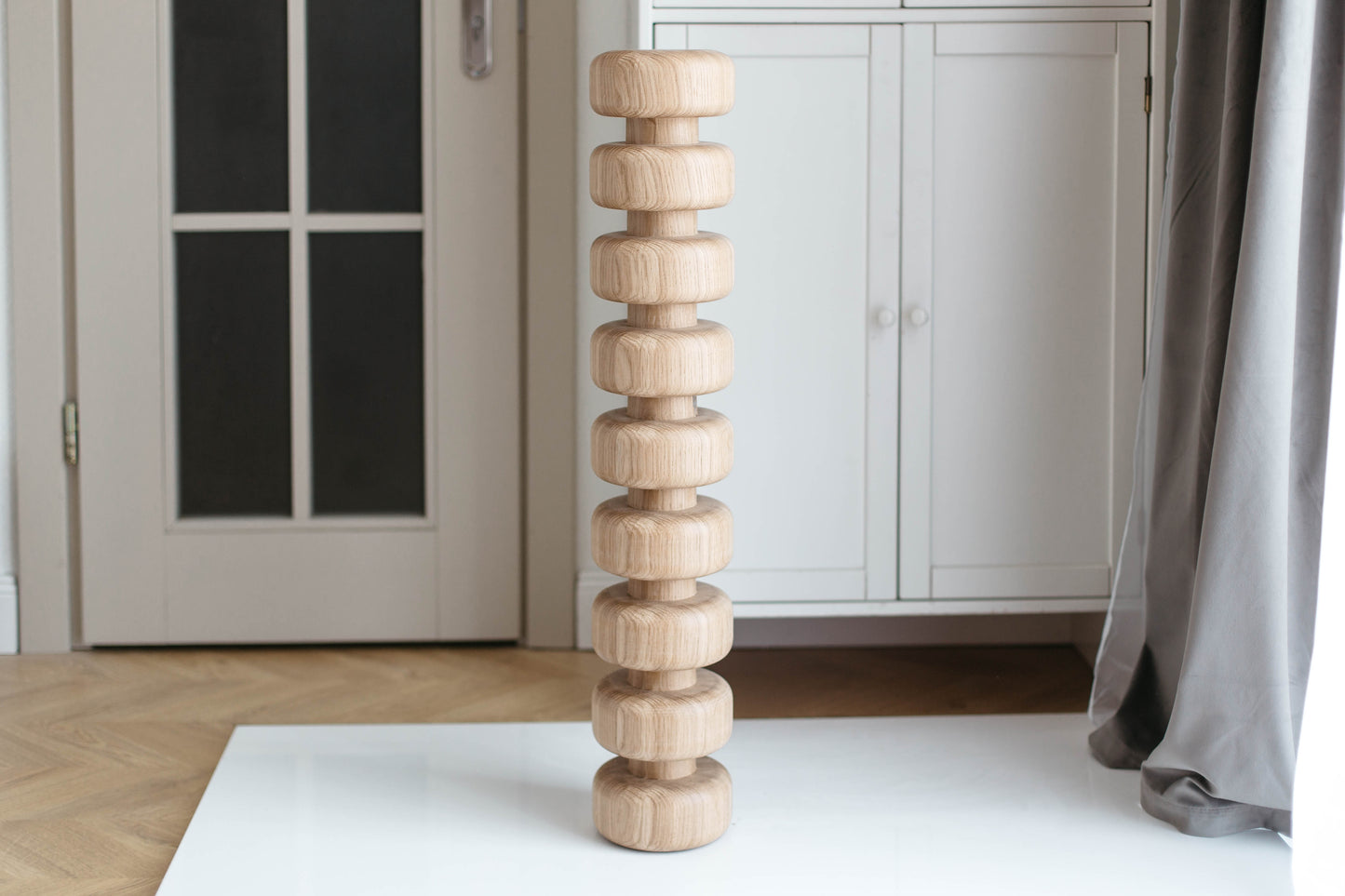 A light natural wood tower of stacked round discs stands on a white surface in a bright room with a white cabinet, gray curtain, and wooden floor in the background.
