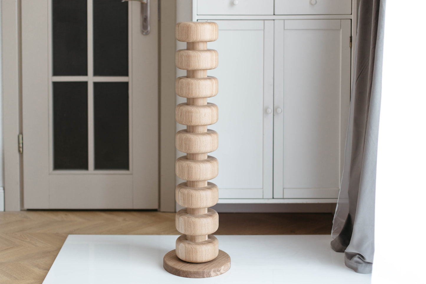 A tall, narrow wooden tower sculpture made of stacked circular rings on a round base, standing on a white table in a bright room with white cabinets and a gray curtain.