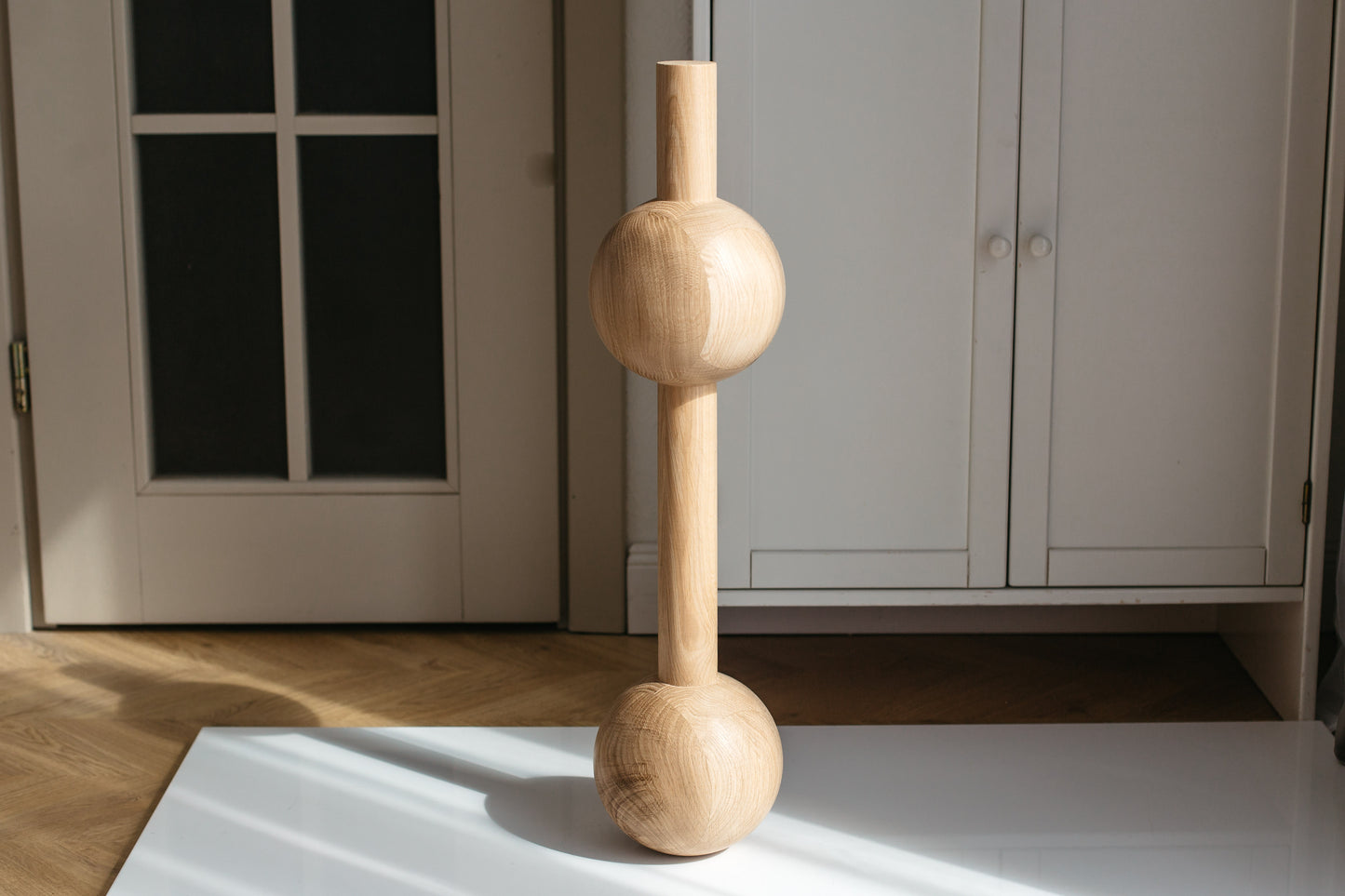 A minimalist wooden sculpture with three stacked spherical and cylindrical forms, standing on a white surface in a bright room with a white cabinet and glass-paneled door in the background.