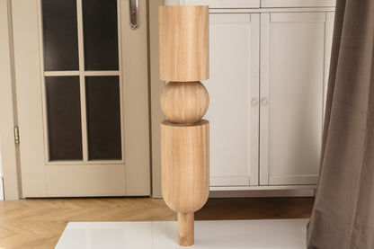 A tall, sculptural wooden floor lamp with a natural light oak finish, featuring stacked cylindrical and spherical segments, shown standing on a white surface in a home interior.