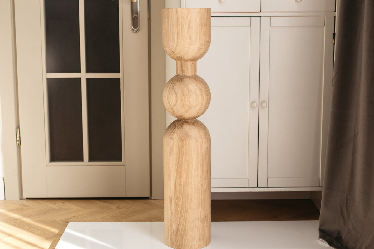 A tall, light natural wood sculptural floor lamp or decorative stand with a flared top, bulbous middle, and rounded base, standing on a white surface in a home interior with cabinets and a door in the background.