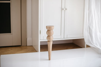 A single wooden furniture leg with a two balls design placed on a white surface with furniture in the background.
