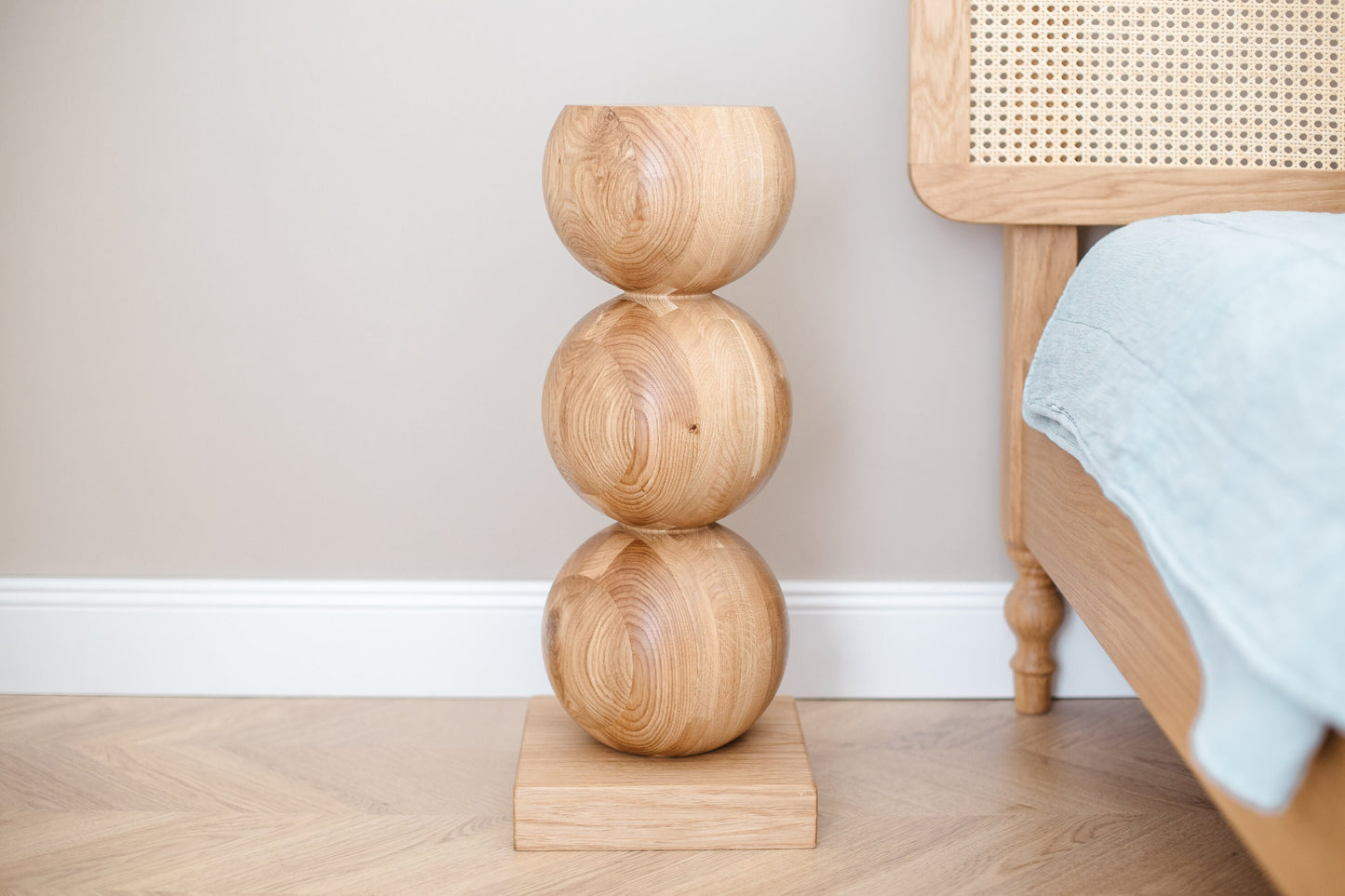 A handcrafted wooden bedside table with a central sphere accent, placed on a floor beside a bed with a blue bedsheet.