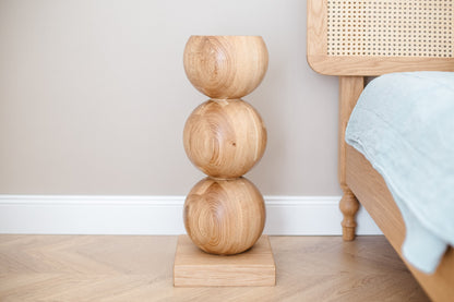 A handcrafted wooden bedside table with a central sphere accent, placed on a floor beside a bed with a blue bedsheet.