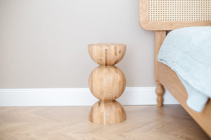 A handturned wooden bedside table with a natural oak finish, featuring a unique wave design, placed on a hardwood floor next to a bed with a blue bedsheet.